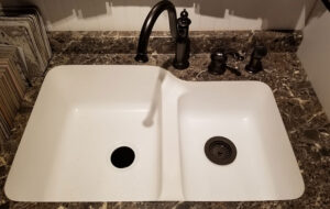 A white acrylic sink from Ray’s Countertop Shop in Glenarm, IL that is installed with a black faucet and brown and white countertop.